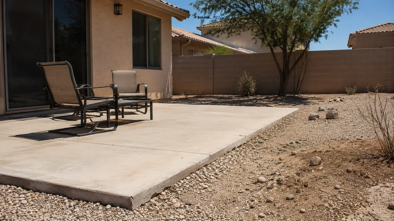 Hot patio edge with exposed hardscape, gravel, sparse planting, and too little softness around the seating area.